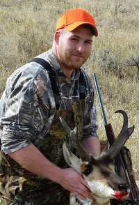 This is an image of a man wearing camouflage hunting gear and an orange cap, posing with a harvested pronghorn antelope. He is holding the animal